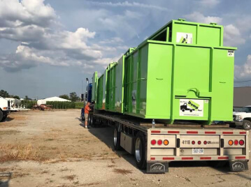 A worker inspecting a flatbed trailer loaded with large green dumpsters from a junk removal franchise.