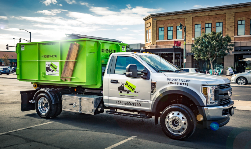 A white truck with a green dumpster attached is parked on a street in front of a commercial building. The truck has the contact number 480-999-1399.