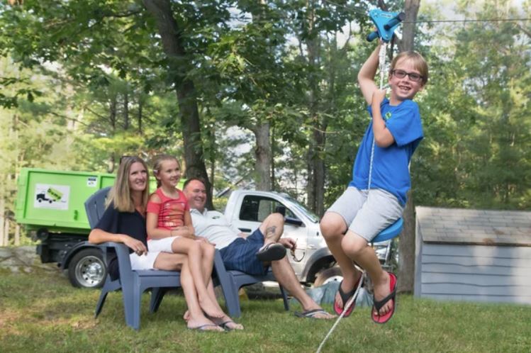 A boy rides a zipline in a backyard while a woman, a girl, and a man sit on chairs, smiling. A shed and a green truck are visible in the background.